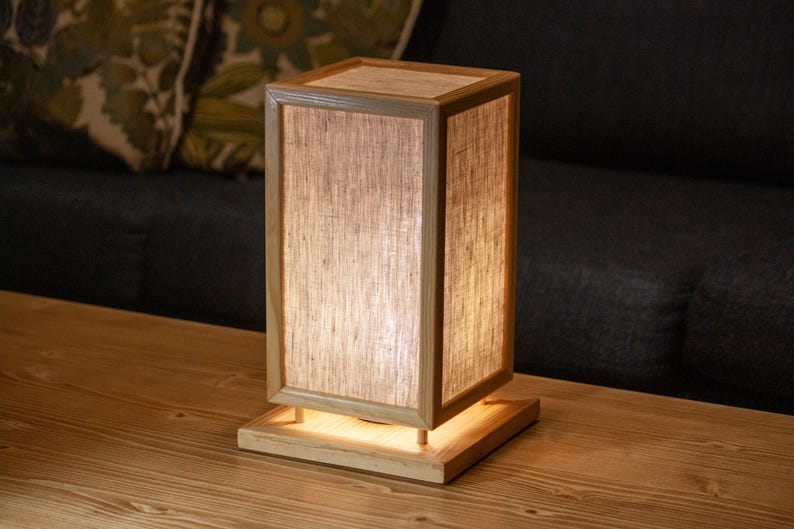 May include: A rectangular wooden table lamp with a light-colored shade. The lamp has a square base and a rectangular frame. The shade is made of a light-colored, translucent material. The lamp is sitting on a wooden table.