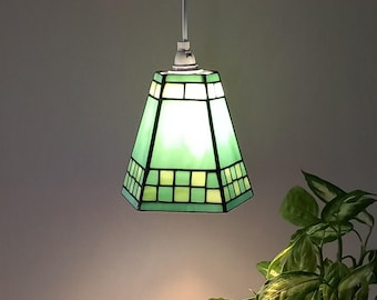 May include: A green and white stained glass pendant light fixture. The lamp has a geometric design with a square pattern at the bottom and a tapered shape. The light is suspended from a white cord.