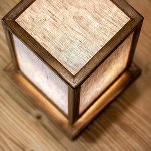May include: A square wooden lamp with a light linen shade. The lamp has a warm, inviting glow, and the wood frame has a rich, dark finish. The lamp sits on a wooden surface.