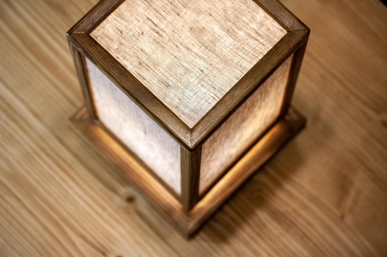 May include: A square wooden lamp with a light linen shade. The lamp has a warm, inviting glow, and the wood frame has a rich, dark finish. The lamp sits on a wooden surface.