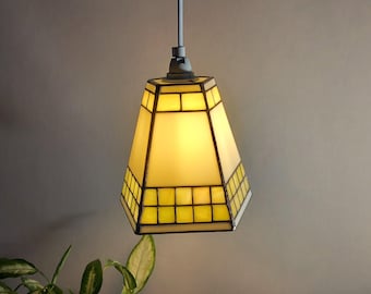 May include: A hanging pendant light with a geometric design. The lamp features a yellow stained glass shade with a black border. The light is suspended from a gray cord and fixture. The lamp is illuminated, casting a warm glow.