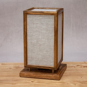 May include: A rectangular wooden lamp with a light gray linen shade. The lamp has a square base and a matching top, with the shade encased in a wooden frame. The lamp is made of wood and linen.