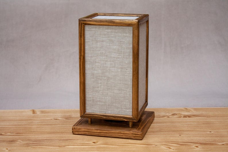May include: A rectangular wooden lamp with a light gray linen shade. The lamp has a square base and a matching top, with the shade encased in a wooden frame. The lamp is made of wood and linen.