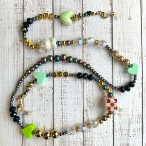 May include: A multi-colored beaded glasses chain with green heart charms, a white bear charm, a gold smiley face charm, and a red and white checkered charm. The chain is made of black, gold, and iridescent beads.