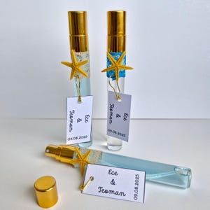 May include: Three clear glass perfume bottles with gold caps and sprayers. Each bottle is decorated with a starfish and a tag that reads "Ece & Teoman" and "09.08.2025". One bottle is lying down with its cap off.