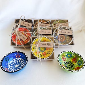 Op de afbeelding: Een verzameling kleine, decoratieve keramische kommen met kleurrijke bloempatronen. Sommige kommen zijn verpakt in transparante dozen met "Thank You" labels vastgebonden met touw. De kommen zijn in blauw, geel, rood en groen.