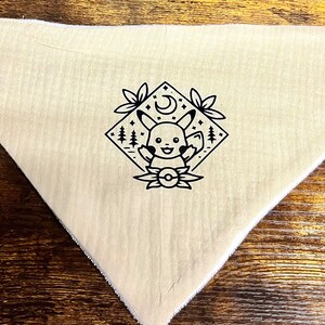 Puede incluir: Un bandana beige con una ilustraci&oacute;n en blanco y negro de Pikachu en un marco de forma de diamante con una luna creciente y estrellas. El bandana tiene dos broches met&aacute;licos en la esquina superior.
