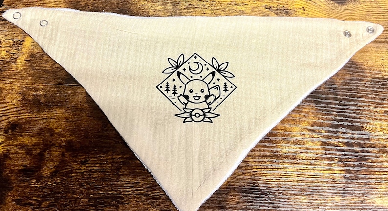 Puede incluir: Un bandana beige con una ilustraci&oacute;n en blanco y negro de Pikachu en un marco de forma de diamante con una luna creciente y estrellas. El bandana tiene dos broches met&aacute;licos en la esquina superior.