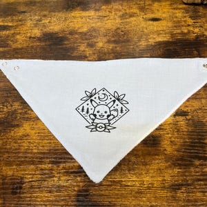 Puede incluir: Bandana de algod&oacute;n blanco con una ilustraci&oacute;n en blanco y negro de un personaje de dibujos animados con una luna creciente y estrellas. El personaje est&aacute; rodeado de hojas y una Pok&eacute; Ball.