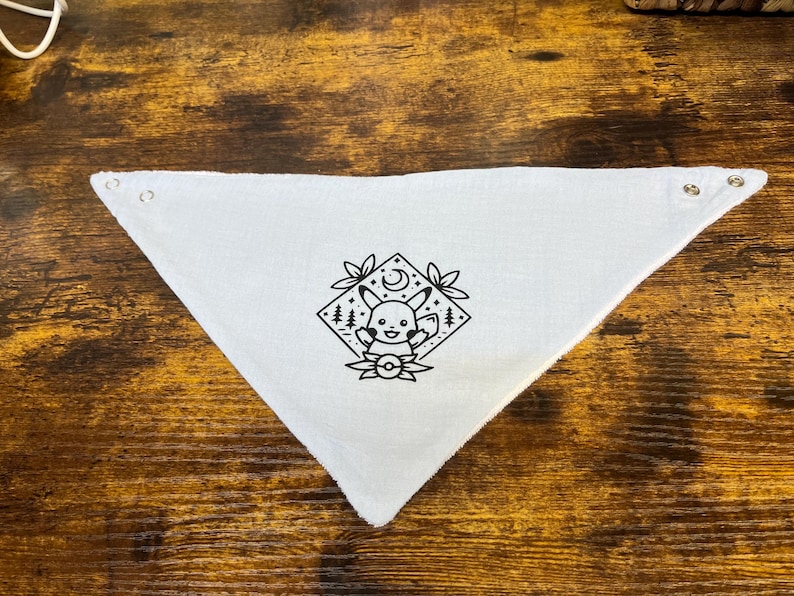 Puede incluir: Bandana de algod&oacute;n blanco con una ilustraci&oacute;n en blanco y negro de un personaje de dibujos animados con una luna creciente y estrellas. El personaje est&aacute; rodeado de hojas y una Pok&eacute; Ball.
