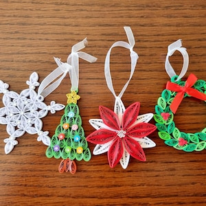 May include: Four handmade Christmas ornaments: a white snowflake, a green Christmas tree, a red and white poinsettia, and a green wreath with a red bow. Each ornament has a white ribbon for hanging.
