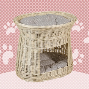 Op de afbeelding: Een wit rieten kattenbed met een grijs en wit gestippeld kussen. Het bed heeft een verhoogd platform en een lager slaapgedeelte.