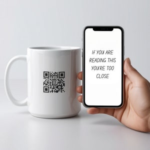 Op de afbeelding: Een witte keramische mok met een zwarte QR-code aan de zijkant. Een hand houdt een smartphone vast met de tekst "Als je dit leest, ben je te dichtbij" op het scherm.