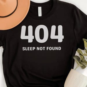 Könnte beinhalten: Schwarzes T-Shirt mit dem weißen Text "404 SLEEP NOT FOUND". Ein beigefarbener Fedora-Hut ist oben links zu sehen. Eine weiße Tasse und Grünzeug befinden sich unten rechts.