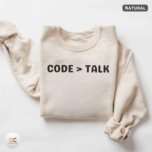 Peut inclure: Un sweat-shirt de couleur naturelle avec le texte "CODE > TALK" imprimé en noir. Le sweat-shirt a un col rond et des manches longues avec les poignets retroussés. Le mot "NATURAL" est imprimé dans un petit encadré en haut à droite.