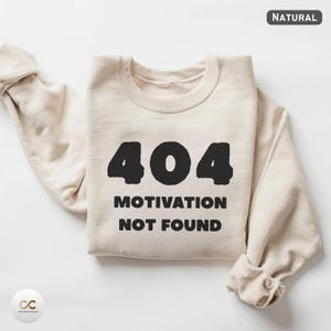 Könnte beinhalten: Beiges Sweatshirt mit dem schwarzen Text "404 MOTIVATION NOT FOUND". Das Sweatshirt ist gefaltet, die Ärmel sind sichtbar. Das Wort "NATURAL" befindet sich in der oberen rechten Ecke.