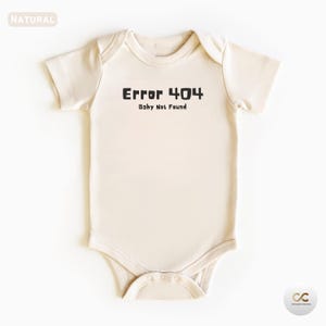 Puede incluir: Un body de bebé color crema con mangas cortas. El texto "Error 404 Baby Not Found" está impreso en negro en la parte delantera. El body tiene un cierre de broche en la parte inferior y un cuello redondo.