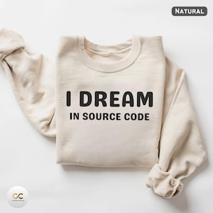 May include: A cream-coloured sweatshirt with the black text "I DREAM IN SOURCE CODE". The sweatshirt has a crew neck and long sleeves, with the sleeves folded up. The word "NATURAL" is in the top right corner.