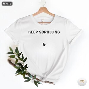May include: White t-shirt with the text "KEEP SCROLLING" above a black cursor icon. The shirt is hanging on a wooden hanger. Green leaves and a branch are placed on the bottom left of the shirt.