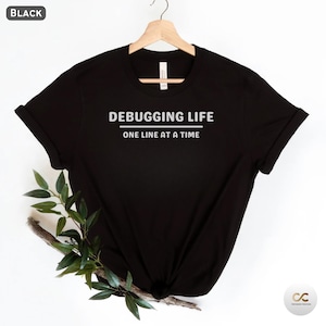 Könnte beinhalten: Schwarzes T-Shirt mit dem weißen Text "DEBUGGING LIFE ONE LINE AT A TIME". Das T-Shirt hängt an einem Holzbügel. Die Ärmel sind hochgekrempelt. Ein Zweig mit Grünzeug befindet sich auf der linken Seite des T-Shirts.