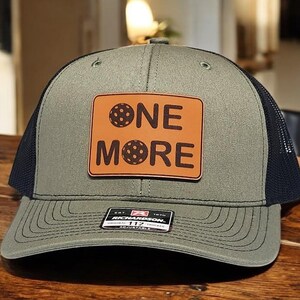 May include: A green and black trucker hat with a brown leather patch that says "ONE MORE" with a black and white disc in the middle of the patch.