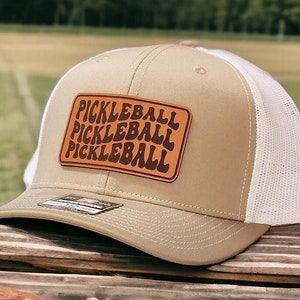 May include: A tan and white trucker hat with a brown leather patch that says "Pickleball" three times in a retro font.