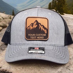 May include: Gray trucker hat with a black mesh back and a brown leather patch featuring a mountain and forest design. The patch includes the text "YOUR CUSTOM TEXT HERE!". The hat has a curved brim and an adjustable strap.