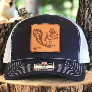 May include: A navy blue and white trucker hat with a brown leather patch featuring a squirrel and the word "NUTS". The hat is made by Richardson 112.