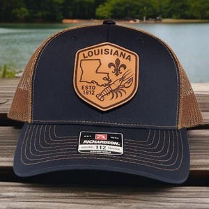 May include: A navy blue and tan trucker hat with a leather patch featuring the state of Louisiana, a fleur-de-lis, and a crawfish. The hat has a curved brim and an adjustable snap closure.