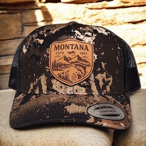 May include: A camouflage-patterned trucker hat with a black mesh back. The front features a leather patch with the word "MONTANA" above an illustration of a mountain, trees, and a fish. The hat has a snapback closure.