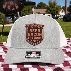 May include: A gray trucker hat with a white mesh back and a brown leather patch. The patch features the words "Beer Bacon Freedom" with an eagle and stars. The hat is sitting on a red and white checkered blanket.