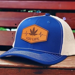 May include: A blue and white trucker hat with a brown leather patch featuring a marijuana leaf and the text "420 LIFE" with a heartbeat graphic.