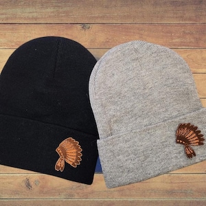 May include: Two knit beanies with a Native American headdress design on the side. One beanie is black and the other is gray.