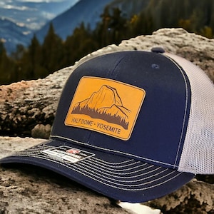 May include: A navy blue trucker hat with a tan leather patch featuring a mountain graphic and the text "HALF DOME - YOSEMITE". The hat has a white mesh back and a curved brim with white stitching. The hat is sitting on a rock.