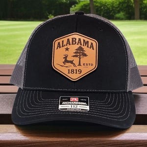 May include: Black trucker hat with a gray mesh back and a curved brim. The front features a brown leather patch with the word "ALABAMA", a star, a deer, a tree, and the year "1819". The hat is adjustable.