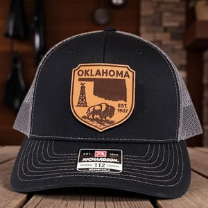 May include: Black and gray trucker hat with a leather patch featuring the state of Oklahoma. The patch includes the word "Oklahoma", a map outline, a bison, and the text "Est. 1907". The hat is adjustable.