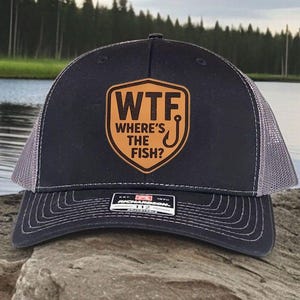 May include: A black trucker hat with a gray mesh back and a curved brim. The hat features a brown leather patch with the text "WTF WHERE'S THE FISH?" and a fish hook graphic. The hat is sitting on a rock.
