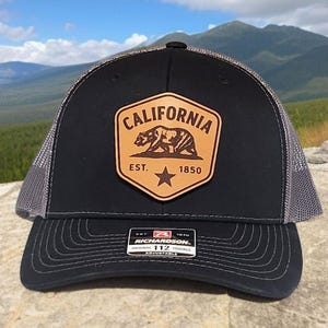 May include: Black trucker hat with a gray mesh back and a curved brim. The front features a brown leather patch with the word "CALIFORNIA" and a bear graphic. The patch also includes the text "EST. 1850" and a star. The hat has a Richardson tag.