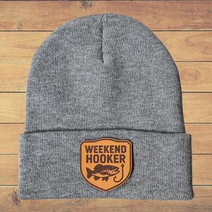 May include: A gray beanie with a brown patch featuring the words "WEEKEND HOOKER" above a fish and hook design. The beanie is made of a soft, knitted material and is displayed on a wooden surface.