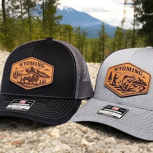 May include: Two trucker hats with a leather patch featuring the word "Wyoming" and outdoor scenes. One hat is black with a bison graphic, and the other is gray with a fisherman graphic. Both have mesh backs and curved brims.