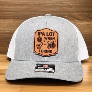 May include: Gray and white trucker hat with a leather patch that reads "IPA LOT WHEN I DRINK" with beer-related graphics. The hat has a curved brim and an adjustable strap.