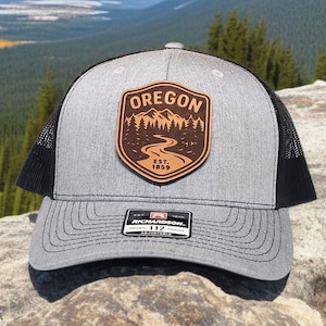 May include: A gray and black trucker hat with a brown leather patch featuring the word "OREGON" above a mountain and river scene. The patch also includes "EST. 1859". The hat has a curved brim and a mesh back.