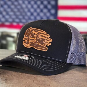 May include: Black and gray trucker hat with a brown leather patch featuring an excavator silhouette against an American flag design.