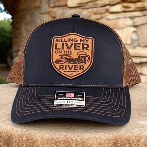 May include: Black and brown trucker hat with a leather patch. The patch features the text "Killing My Liver On The River" with a boat and beer mug graphic. The hat is adjustable.