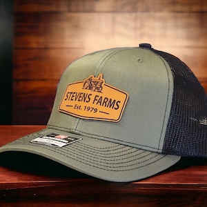 May include: A green and black trucker hat with a curved brim. The hat features a leather patch with the text "STEVENS FARMS" and "Est. 1979" along with a tractor graphic. The hat is on a wooden surface.