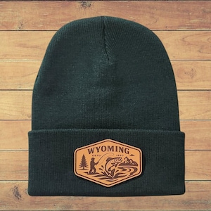 May include: A dark green beanie with a brown leather patch. The patch features the word "WYOMING ESTO 1890" and an illustration of a person fishing, a fish jumping, trees, and mountains.