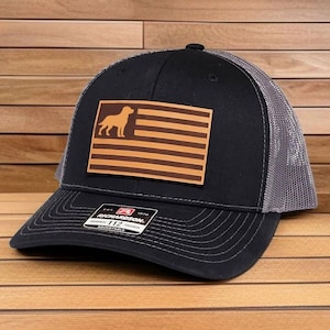 May include: A black trucker hat with a grey mesh back and a rectangular brown leather patch on the front. The patch features a silhouette of a dog standing on the left side of horizontal stripes resembling a flag. A sticker is on the brim.