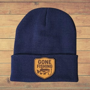 May include: A navy blue knit beanie with a brown leather patch. The patch features the words "GONE FISHING" above a fish and a fishing hook. The beanie is displayed on a wooden surface.