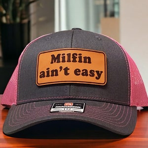 May include: A black and pink trucker hat with a brown leather patch that says "Milfin ain't easy".