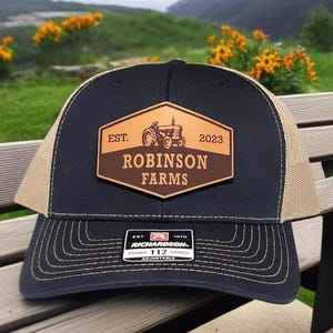May include: A navy blue and tan trucker hat with a brown leather patch. The patch features a tractor graphic and the text "EST. 2023 ROBINSON FARMS." The hat has a curved brim and an adjustable snap closure.